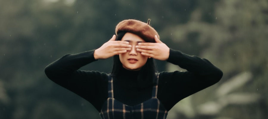 woman with black hair and brown woollen hat holding her hands over both eyes against blurry nature background