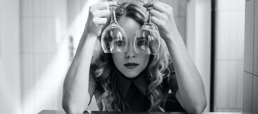 black and white portrait of young woman holding empty wine glasses upside down over her eyes while resting her elbows on the table