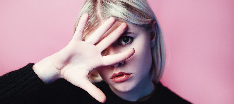 woman with blond hair in black shirt covering one eye with one hand against pink background