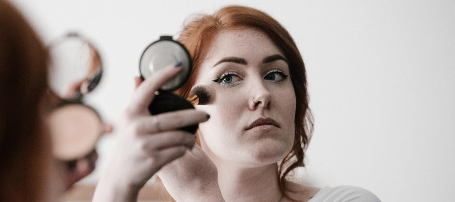 woman with red hair applying makeup while looking in a small round mirror