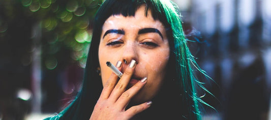 woman with green hair holding cigarette between lips against blurry outdoor background
