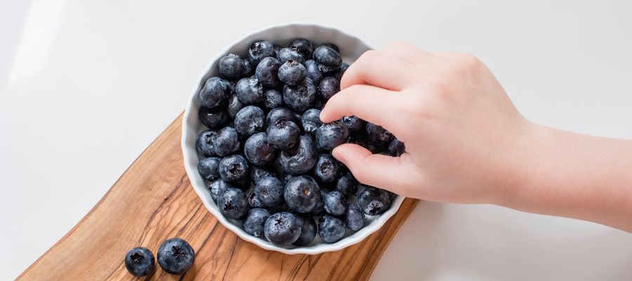hand picking blueberries from a bowl that sits on wooden cutter against white background