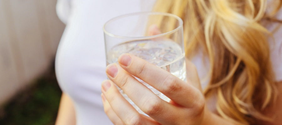 glass of water in woman's hand with her white t-shirt and blond hair in the background