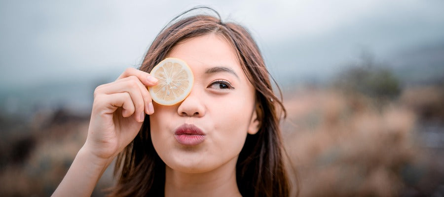 portrait of woman holding a lemon slice over one eye and pouting lips with the other eye open and blurry nature background