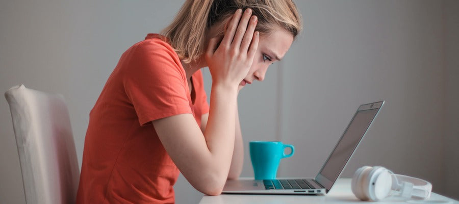 Woman in red t-shirt hunched over computer holding head in her hands with worried expression