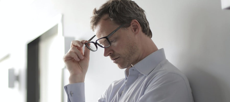 man in a shirt taking off black eyeglasses while looking down with half-closed eyes against blurry white background