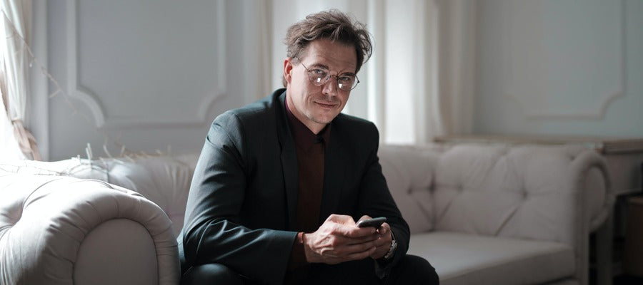man with glasses in dark business suit sitting on couch smiling while buttoning phone