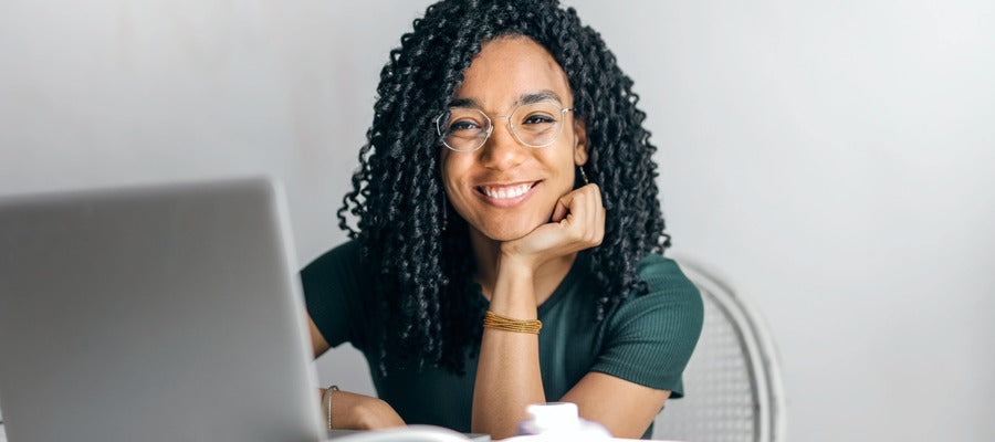 woman in green shirt with glasses and with curly Afro hairstyle smiling next to computer while holding hand under chin