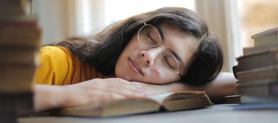 woman with glasses lying with eyes closed on open book between stacks of books having tired eyes