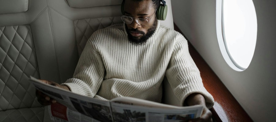 African American man in white sweater with glasses and headphones reading newspaper in business class airplane seat