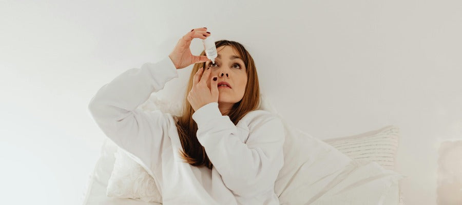 woman in white room putting eye drops in one eye while sitting on the couch