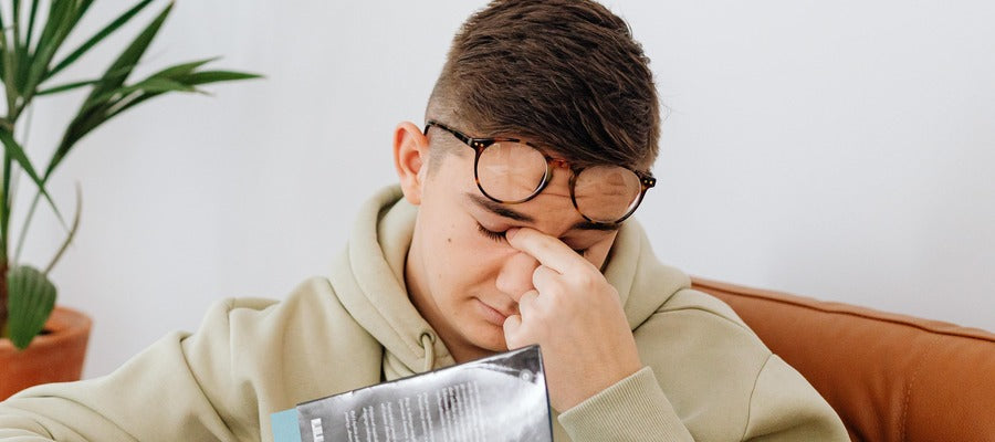 young man wearing eyeglasses over forehead rubbing eyes while holding book with white wall and plant in background