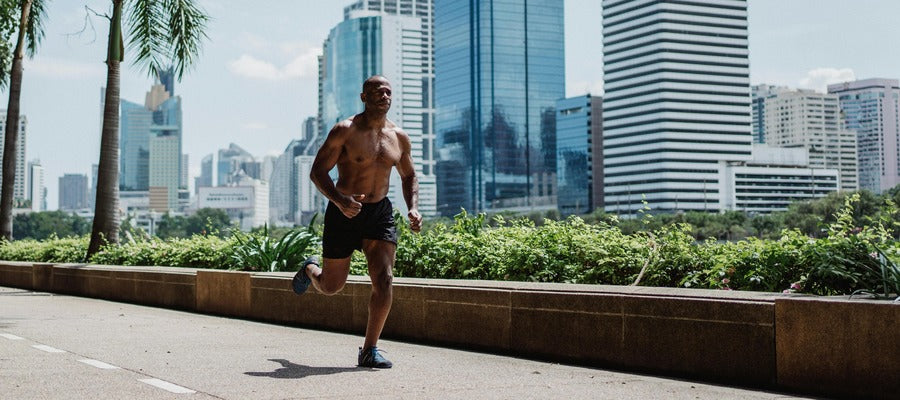 African American man running on the street bare chested on sunny day in black shorts with greenery, skyscrapers, and palms in background