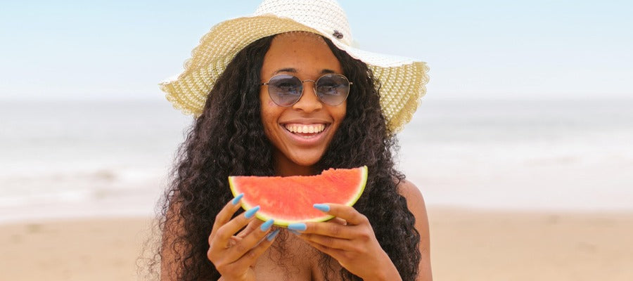 portrait of woman at the beach with sunglasses and large hat smiling and holding watermelon slice with blurry sand and sea in the background