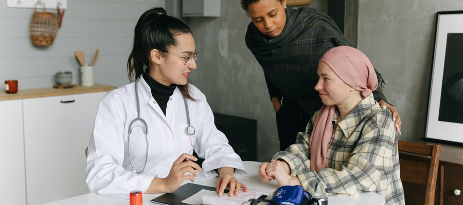 female doctor in white coat sitting at table with woman with pink scarf over head who is undergoing cancer treatment while another woman leans over them in the background