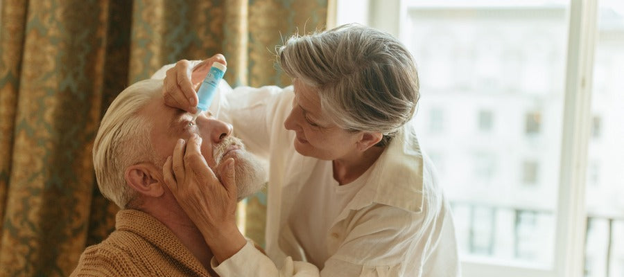 woman with silver hair putting eye drops into seated old man's eye
